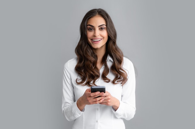 Satisfied happy smiling business woman types text message on cell phone enjoys online communication types feedback isolated on gray studio wall