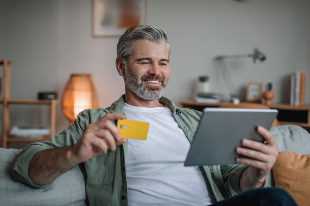 Satisfied mature european man with beard shopping online hold tablet show credit card in room interior