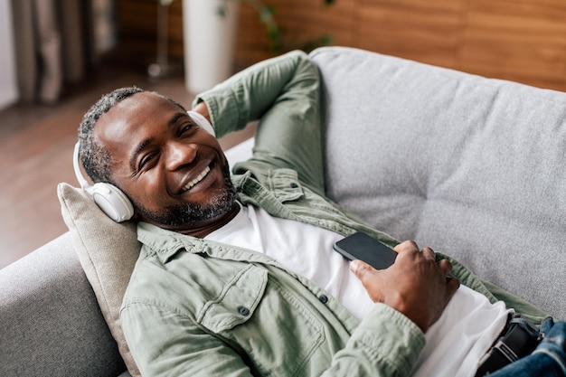 Satisfied middle aged african american man in casual in wireless headphones enjoys favorite music with phone