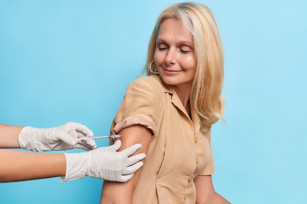 Satisfied middle aged woman feels protected after making inoculation against coronavirus poses against blue wall