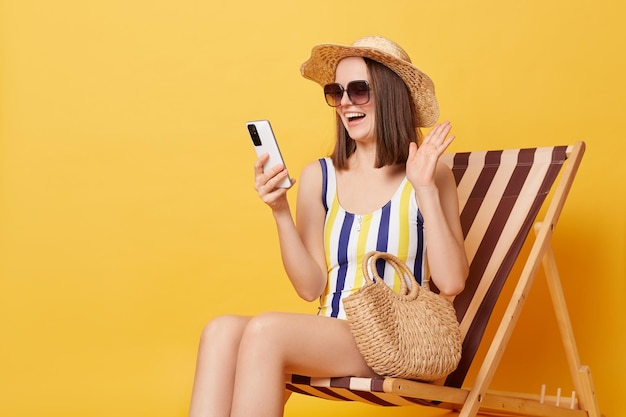 Satisfied positive woman wearing striped swimwear and hat sitting on deck hair resting at resort having video call waving hand saying hello