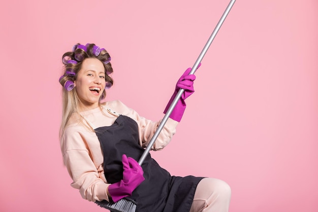 Satisfied smiling woman with long blonde hair curled rollers on head has protective rubber cleaning gloves on hands holds a broom pretending to play guitar fooling around