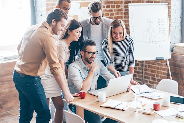 Satisfied with new plan. Group of six cheerful young people looking at laptop with smile while leaning to the table in office