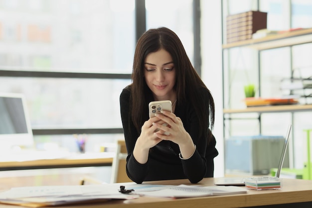 Satisfied woman sends messages to boyfriend via smartphone sitting at table in office brunette