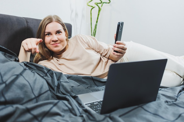 Satisfied young woman with phone continuing work on laptop Woman sitting with computer in bed studying at home distance learning and remote freelance work concept