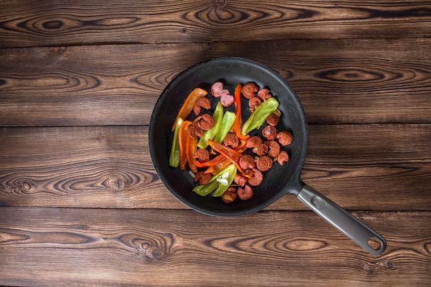 Sausages on the grill pan on the wooden background. Top view. Frying pan with fried sausage and vegetables on a rustic wooden table.