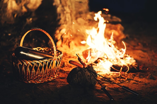 Sausages on skewers and a thermos in the basket against the fire at night.