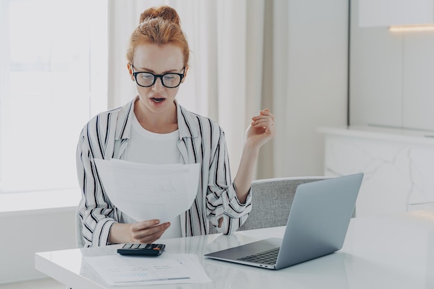 Savings finances concept Shocked ginger woman focused at document studies bills reads banking paper