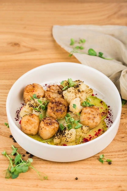 Scallop baked with cauliflower in a plate with sauce on a light wooden surface close-up.