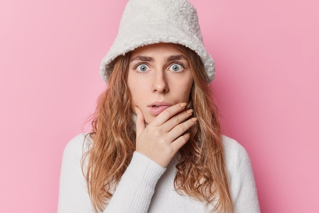 Scared fearful woman holds chin stares with eyes full of amazement notices horrible thing wears white hat and turtleneck isolated over pink background. Frightened female model afraids of something