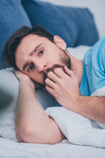 Scared man lying in bed looking at camera and covering mouth with hand