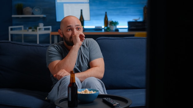 Scared man watching horror film on tv eating popcorn sitting on sofa