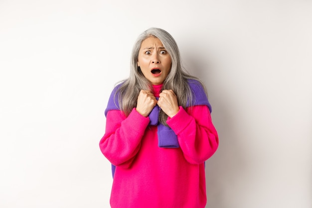 Scared and shocked asian grandmother gasping and staring frustrated at camera, standing against white background