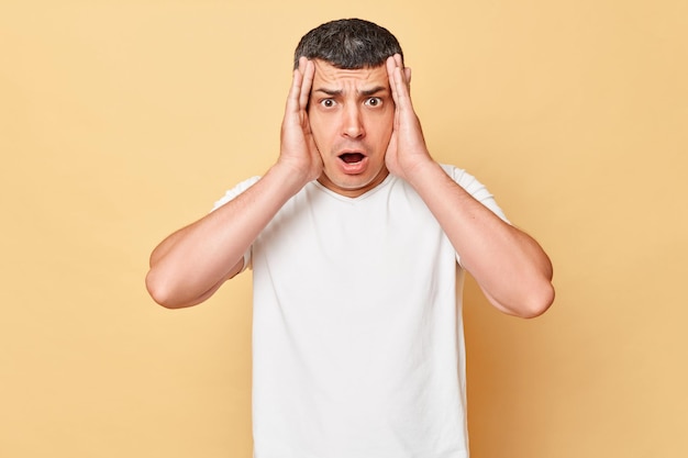 Scared shocked panicked man wearing white casual tshirt standing isolated over beige background looking at camera with big full of fear eyes raised arms saying omg