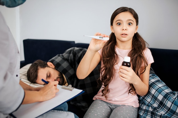 Scared small girl holding thermometer and bottle of syrup in hands. Female doctor sit besides her and write. Sick young man lying on bed and sleeping.