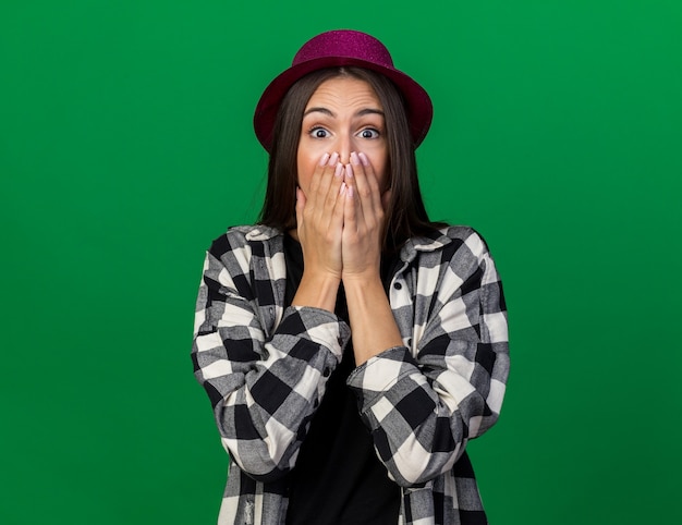 Scared young beautiful girl wearing party hat covered mouth with hand isolated on green wall