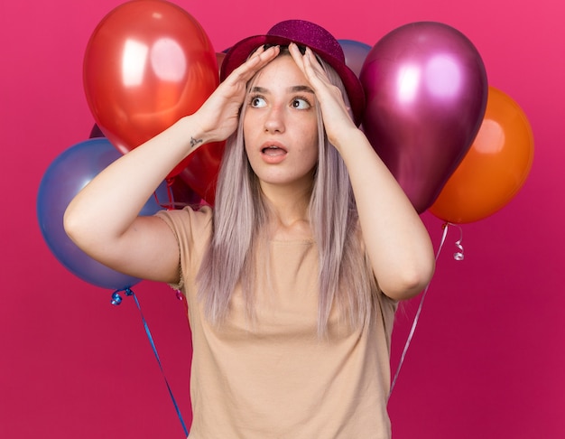 Scared young beautiful girl wearing party hat standing in front balloons putting hands on rorehead isolated on pink wall