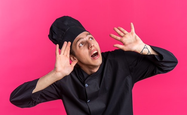 Scared young blonde male cook in chef uniform and cap looking up showing empty hands 