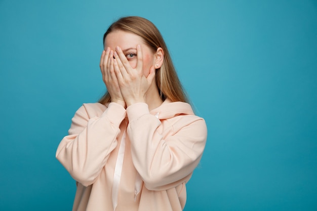 Scared young blonde woman covering face with hands peeking at camera between fingers 