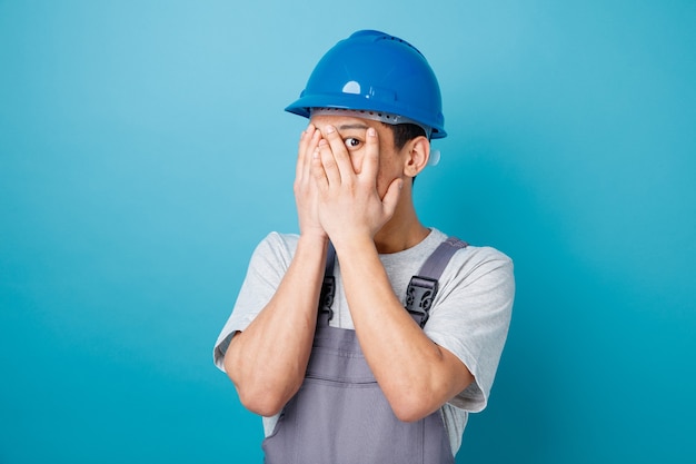 Scared young construction worker wearing safety helmet and uniform covering face with hands peeking at camera between fingers 