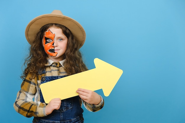 Scary bright alive little girl child with Halloween makeup mask showing aside with yellow arrow, wears hat and shirt, model over blue studio background wall with copy space for promotional content