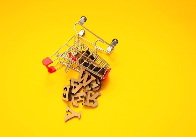 Scattered wooden letters of the English alphabet from a miniature shopping trolley on a yellow background, close-up. Copywriting and education concept. Copy space.