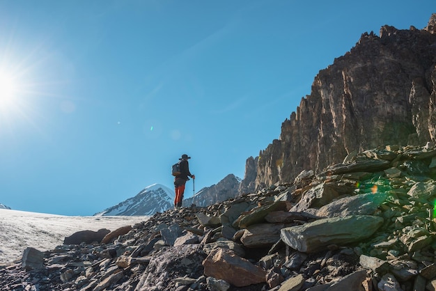 Scenic alpine landscape with silhouette of hiker with trekking poles against large sharp rocks and snow mountain peak in sunlight Man with backpack in high mountains under blue sky in sunny day