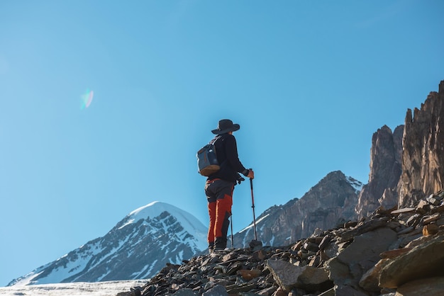 Scenic alpine landscape with silhouette of hiker with trekking poles against large sharp rocks and snow mountain peak in sunlight Man with backpack in high mountains under blue sky in sunny day