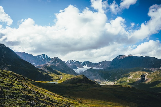 Scenic green blue alpine landscape with mountain lake in highland valley in sunlight and big glacier under cloudy sky. Shadow of clouds on green mountain valley. Clouds shadow on rocks and hills.