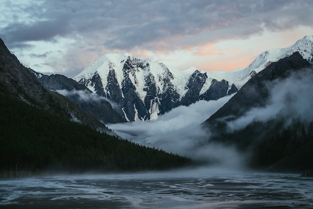 Scenic landscape with great snowy mountains on sunset and dense low clouds in mountain valley with forest silhouette. Atmospheric scenery with high snow-capped mountain on sunrise and thick low clouds