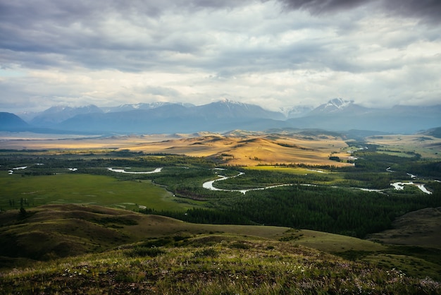 Scenic landscape with vast plateau with mountain river and forest in sunlight on background of snowy mountain ridge under cloudy sky. Green mountain valley in sunshine and mountain range on horizon.