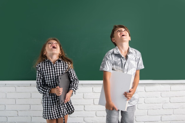 School children hold book with surprising expression against blackboard. School kids friends.