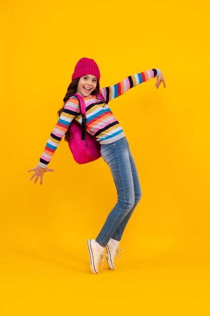 School children in winter hat and sweater with school bag on isolated yellow studio background