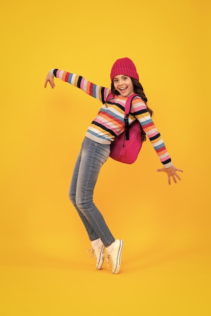 School children in winter hat and sweater with school bag on isolated yellow studio background