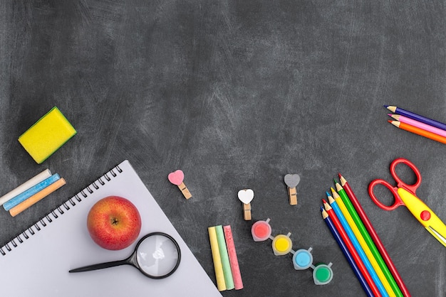 A school desk with a book, pencils, pens, and a red apple on it.