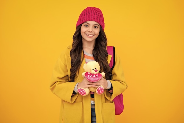 School girl in winter hat hold toy School children with favorite toys on yellow isolated background Childhood concept Happy teenager positive and smiling emotions of teen girl