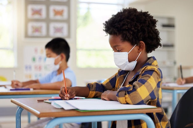 School student in class during covid pandemic for learning education and study with mask for safety protection and protocol Little kindergarten preschool or elementary kid writing in book at desk