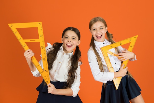 School students learning geometry. Kids school uniform on orange background. Pupil cute girls with big rulers. Geometry school subject. Drawing with ruler chalkboard. Education and school concept.