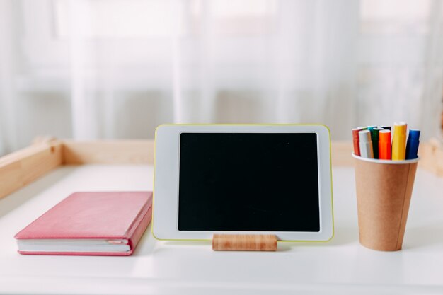 School supplies on the white table. White tablet, pink notebook, marker pens on the table.