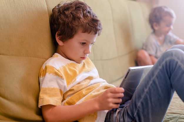 schoolboy 7 years old lies on green couch and enthusiastically plays a game on tablet. High quality photo