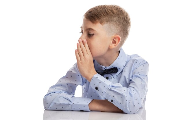 A schoolboy boy in a blue shirt speaks in a whisper with his hand to his mouth. Isolated on white wall. Space for text.