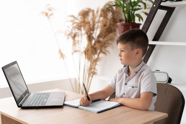 Schoolboy boy studying at home doing homework with a laptop writing down the task in a notebook or notepad junior elementary school student studies at home Distance learning and education concept