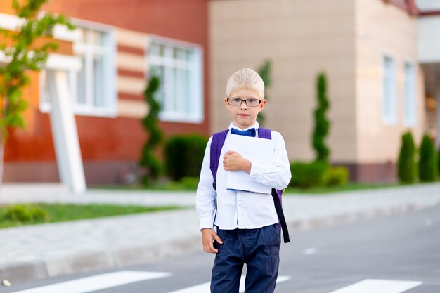 A schoolboy boy with blond glasses with a backpack and a white book is standing at the school