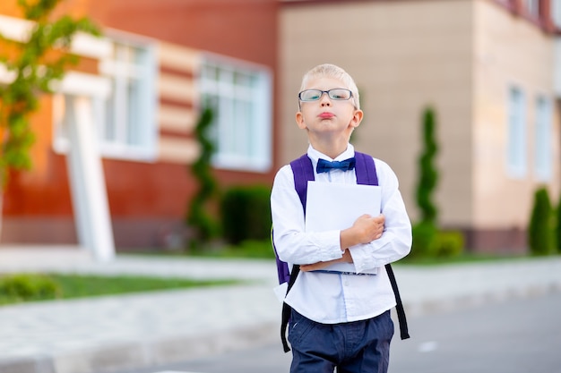 A schoolboy boy with blond glasses with a backpack and a white book is standing at the school