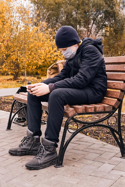 Schoolboy in a protective mask sits on a bench and holds a smartphone New normal