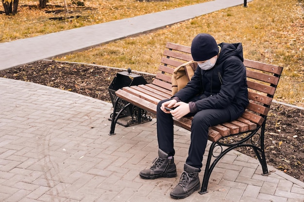 Schoolboy in a protective mask sits on a bench and holds a smartphone New normal