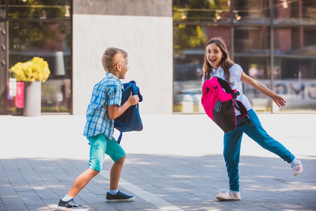 Schoolboy and schoolgirl having fun spending free time outdoors after school lessons Laughing girl with her backpack on chest blond boy holding backpack in front of him