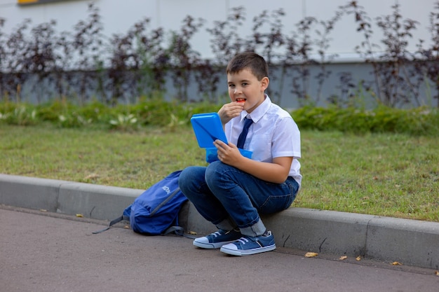 Schoolboy in a white shirt with a blue tie, holds a blue lunch box and eats a piece of apple