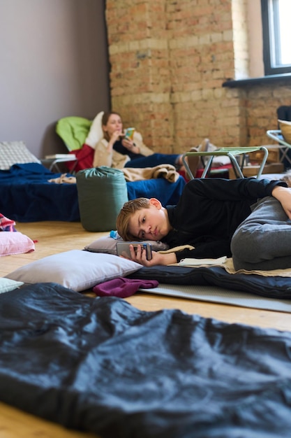 Schoolboy with mobile phone resting on sleeping place on the floor and texting