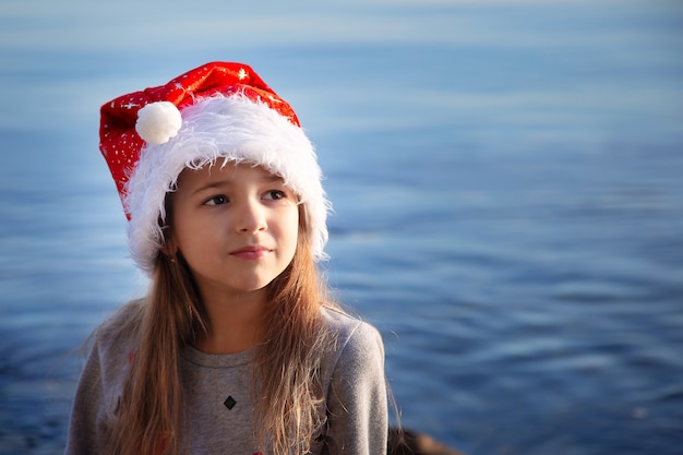 A schoolgirl girl in a New Year's hat sits on the seashore and dreams